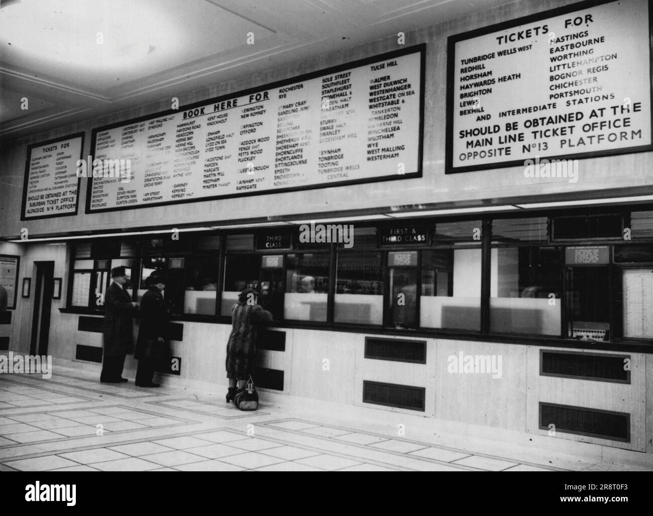 Schnellere Tickets in Victoria -- Ein Blick auf die wiederaufgebaute Buchungshalle des östlichen Abschnitts in Victoria Station (London), wo Sie das Ticketbüro mit Glasfront und die Passagiere, die Tickets kaufen, sehen. Die neue Halle, die bis heute eröffnet wurde, ersetzt die, die während des Krieges schwer beschädigt wurde. Es werden Tickets für den Großteil von Kent serviert. Zentral beheizt und mit fluoreszierendem Licht beleuchtet - in mittelgroßen Weiß- und Goldtönen für eine warme und komfortable Atmosphäre - es enthält Bücherregal, Telefone und Fahrplanständer. Hinter den Kulissen gibt es neue Gestelle für die Buchhalter, die für die schnellen ISSU entworfen wurden… Stockfoto