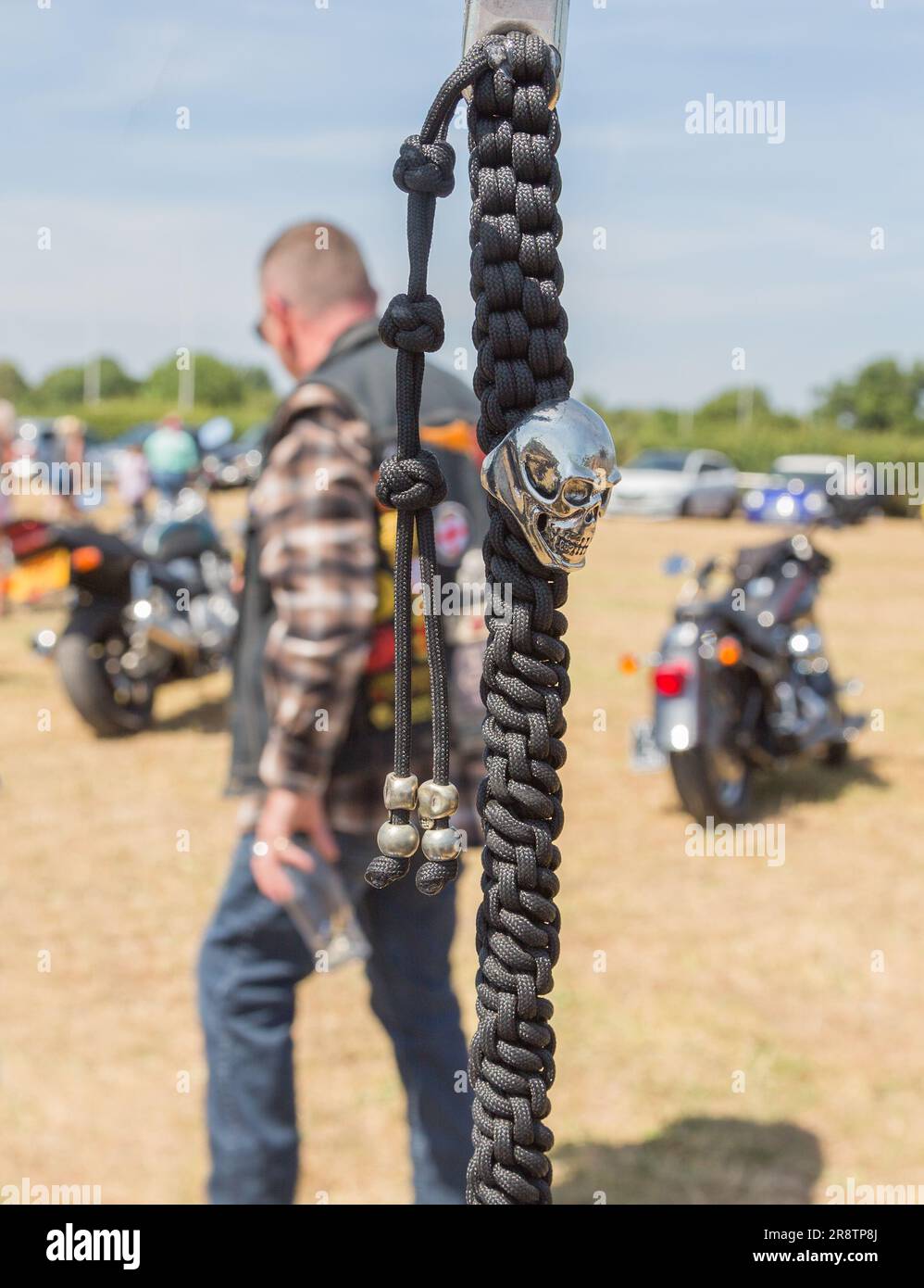 Ein silberner Schädel auf einer schwarzen geflochtenen Kette, die an einem Motorradlenker hängt. Ein Biker, der vorbeiläuft und ein Bierglas hält, verleiht der Szene ein ausgefallenes Gefühl. Stockfoto