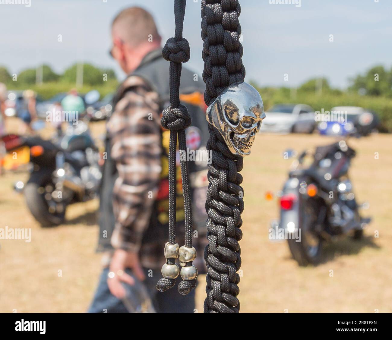 Ein silberner Schädel auf einer schwarzen geflochtenen Kette, die an einem Motorradlenker hängt. Ein Biker, der vorbeiläuft und ein Bierglas hält, verleiht der Szene ein ausgefallenes Gefühl. Stockfoto