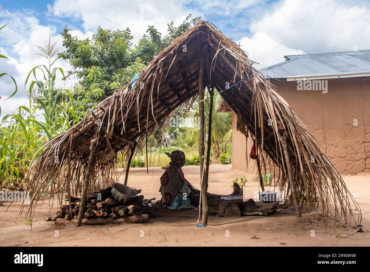 Eine alte Dame aus dem Makonde-Stamm sitzt in der primitiven Küche und bereitet lokale traditionelle Mahlzeit zu. Einfacher Metalltopf auf Feuer zwischen Steinen Stockfoto