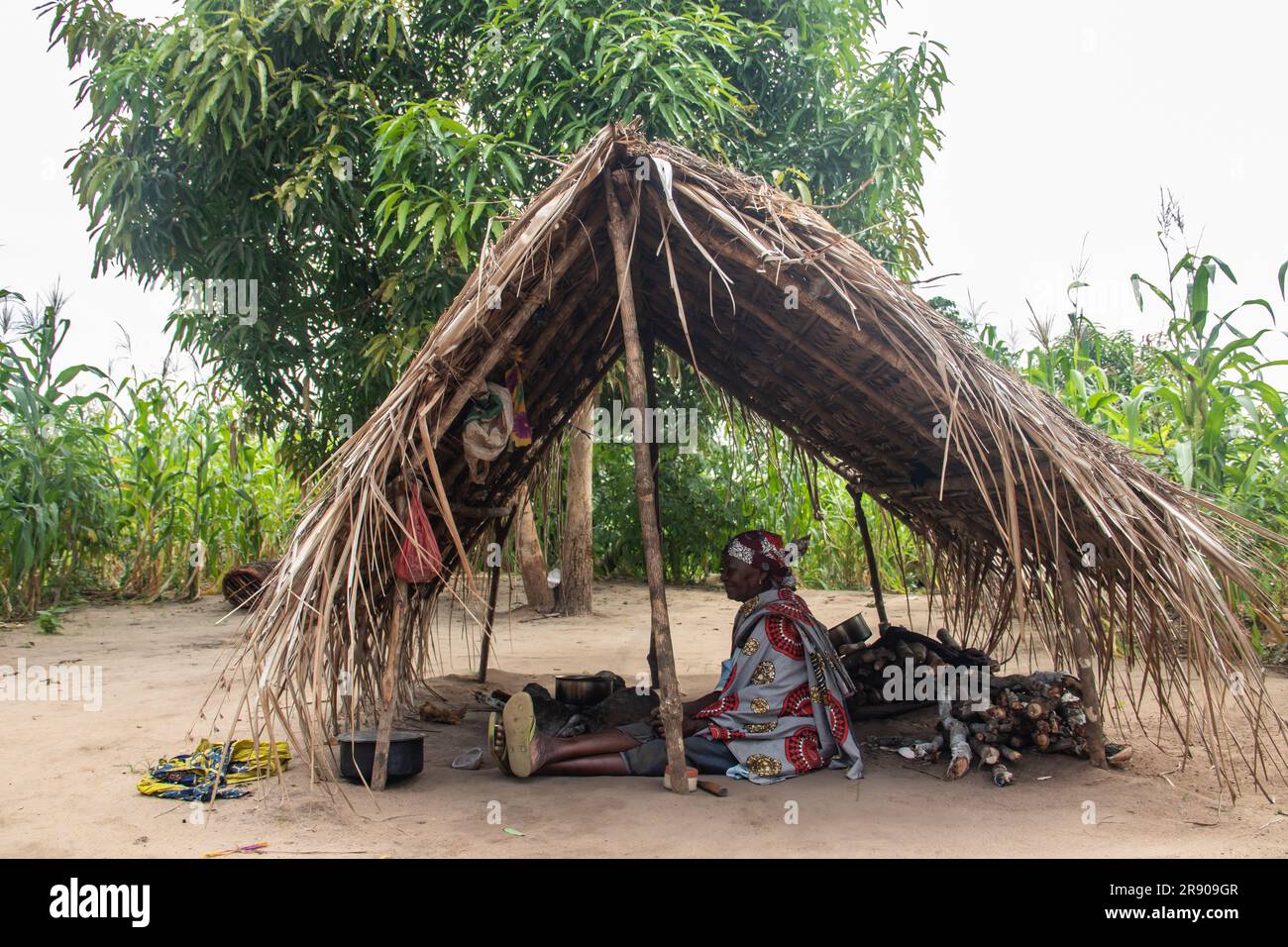 Eine alte Dame aus dem Makonde-Stamm sitzt in der primitiven Küche und bereitet lokale traditionelle Mahlzeit zu. Einfacher Metalltopf auf Feuer zwischen Steinen Stockfoto