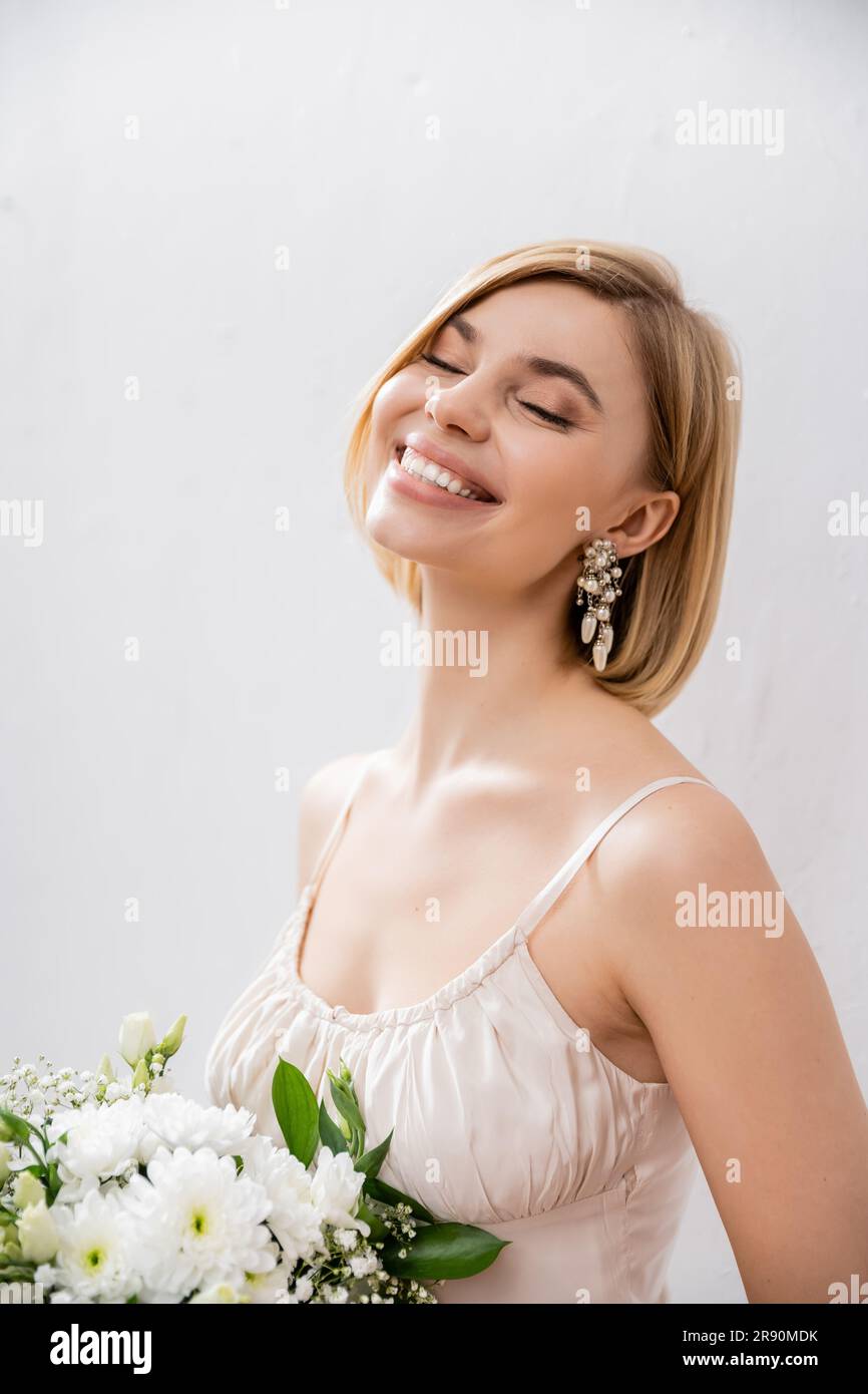 Schöne und blonde Braut im Hochzeitskleid mit einem Strauß auf grauem Hintergrund, Freude, weiße Blumen, Hochzeitszubehör, Glück, Besonderer Anlass, F. Stockfoto