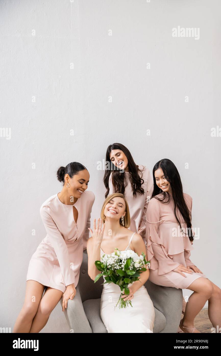 Hochzeitsfotos, Vielfalt, vier Frauen, fröhliche Braut mit Blumenstrauß, die ihren Verlobungsring in der Nähe der Brautjungfern zeigt, Hochzeitstag, auf dem Sessel sitzt, Stockfoto