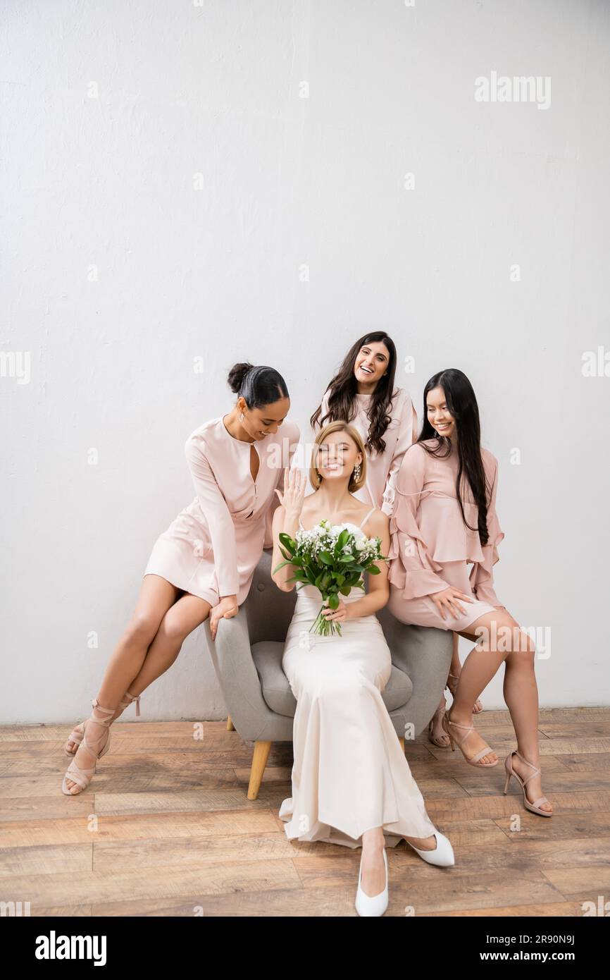 Hochzeitsfotografie, kulturelle Vielfalt, vier Frauen, fröhliche Braut mit Blumenstrauß, die ihren Verlobungsring in der Nähe der Brautjungfern zeigt, Hochzeitstag, auf einem sitzt Stockfoto