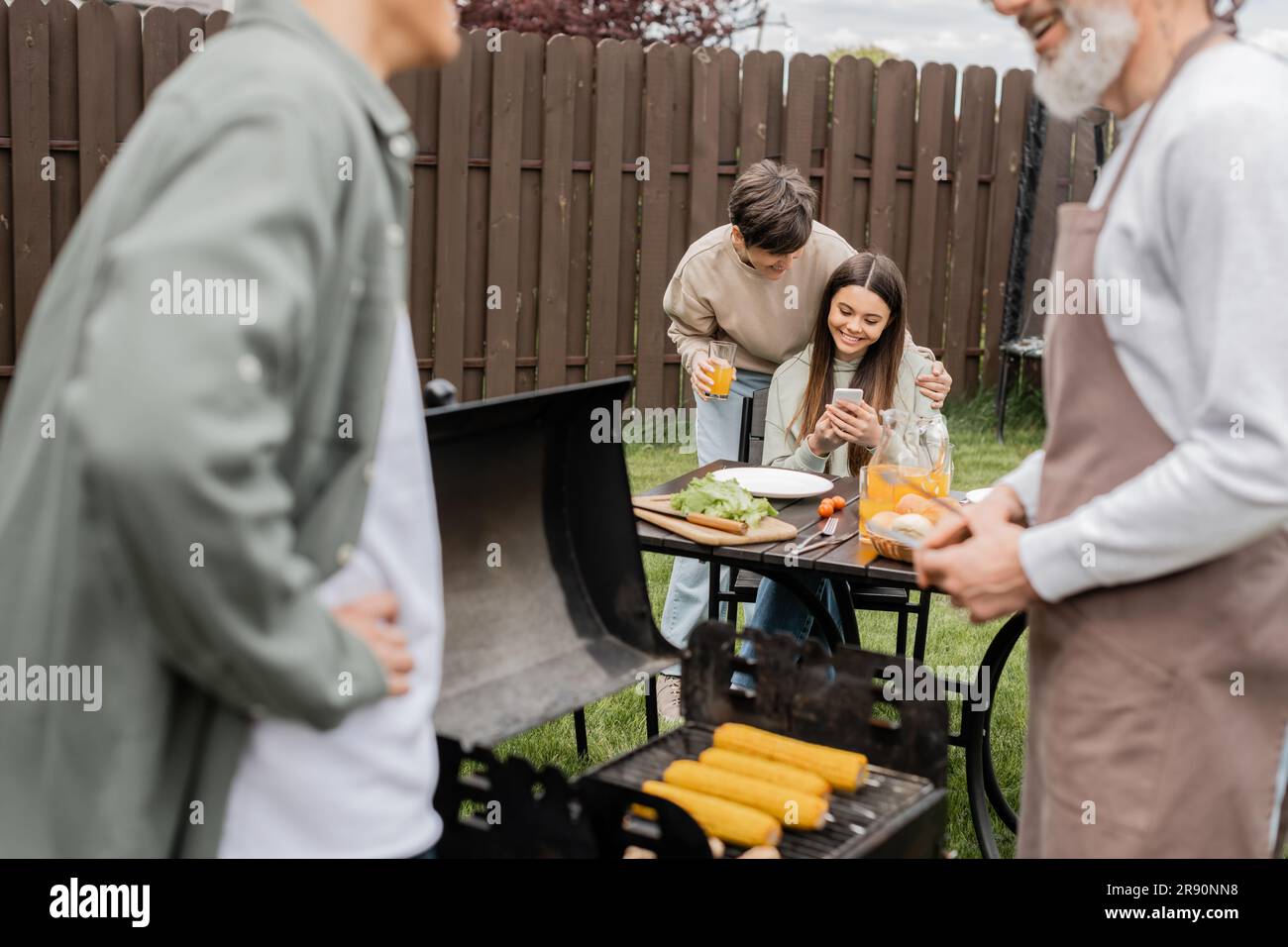 Ein fröhliches Teenager-Mädchen, das dem jungen erwachsenen Bruder, digitalen Zeitalter, dem Vater, der Essen auf dem grill zubereitet, einer Barbecue-Party, den Eltern, etwas auf dem Smartphone zeigt Stockfoto