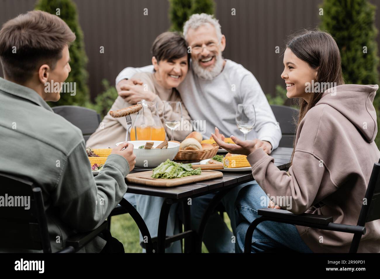 Lächelnde Kinder, die sich unterhalten, während sie neben dem Sommeressen sitzen, und verschwommene Eltern mittleren Alters im Hintergrund während der Elterntagsfeier im Stockfoto