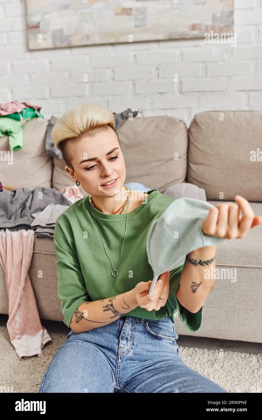 garderobe, die aufblüht, junge und fröhliche Frau mit Tattoo und trendiger Haarschutzkappe neben dem Sofa mit Kleidung im Wohnzimmer, nachhaltig Stockfoto