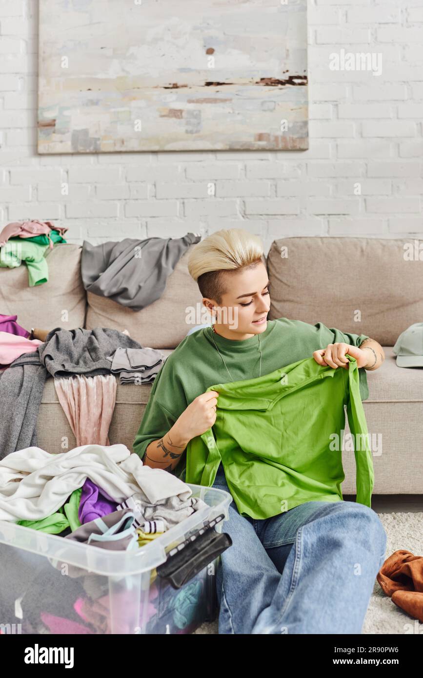 Junge Frau mit trendigen Haaren und Tätowierungen, die grüne Kleidung in der Nähe eines Plastikbehälters und einer Couch mit Kleidung hält, sortiert, bewusst entkleidet, su Stockfoto