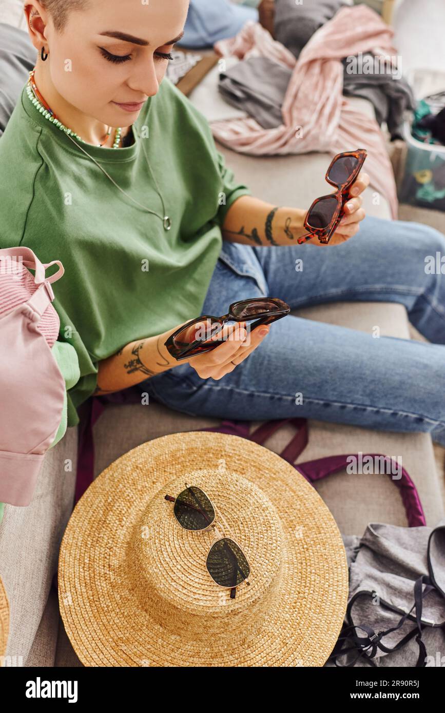 Blick von oben auf eine stilvolle und tätowierte Frau, die eine Sonnenbrille neben einem Strohhut und Kleidungsstücken auf dem Sofa hält, Kleidung sortiert, nachhaltige Livi Stockfoto
