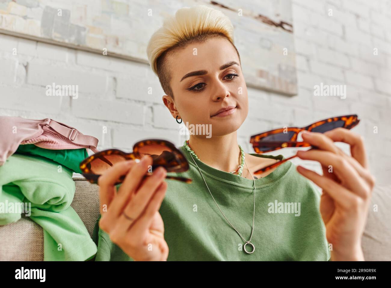 Junge und zufriedene Frau mit trendigen Haaren und Tattoo, die sich eine modische Sonnenbrille in der Nähe der Kleidung auf der Couch zuhause ansieht, vorgeliebte Gegenstände sortiert, Su Stockfoto
