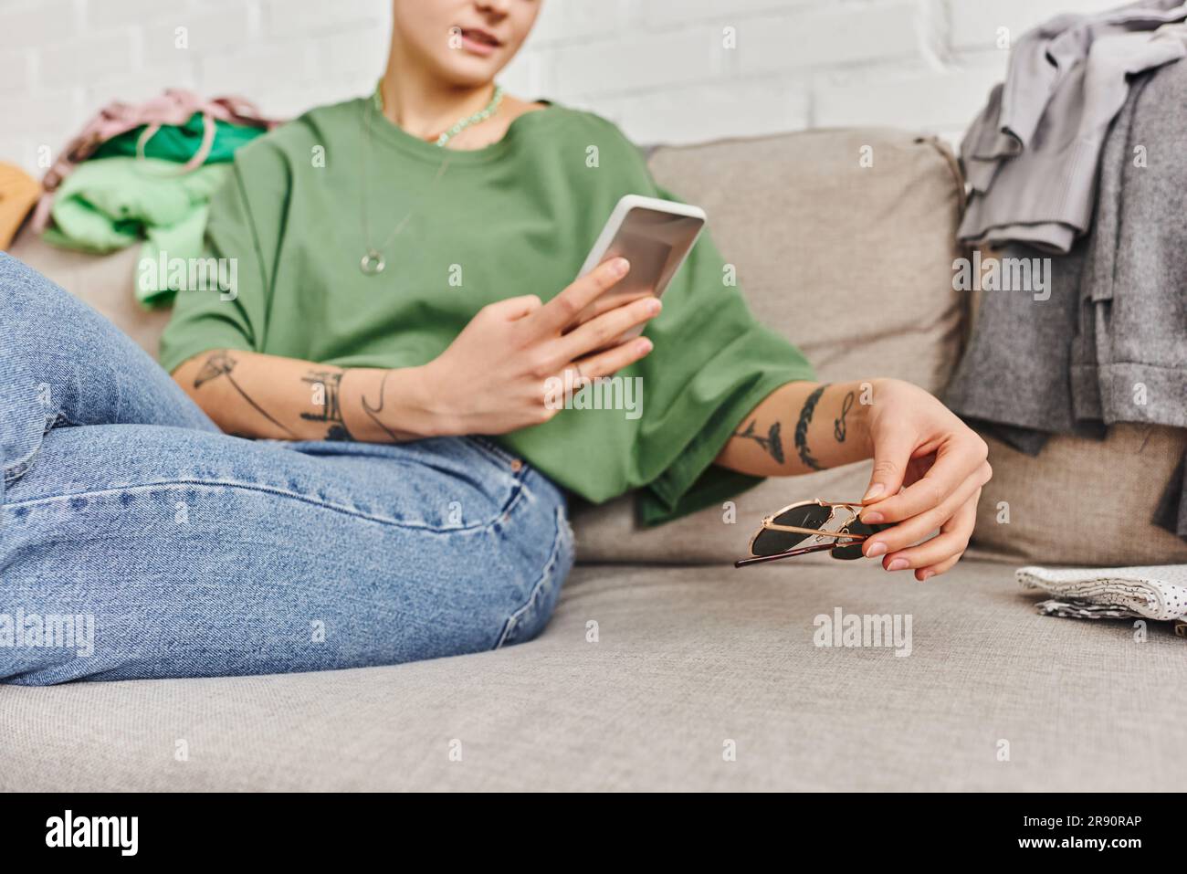Freigestellte Ansicht einer jungen und tätowierten Frau in legerer Kleidung, die zu Hause auf dem Sofa sitzt und mit dem Smartphone eine Sonnenbrille fotografiert, um online zu tauschen Stockfoto