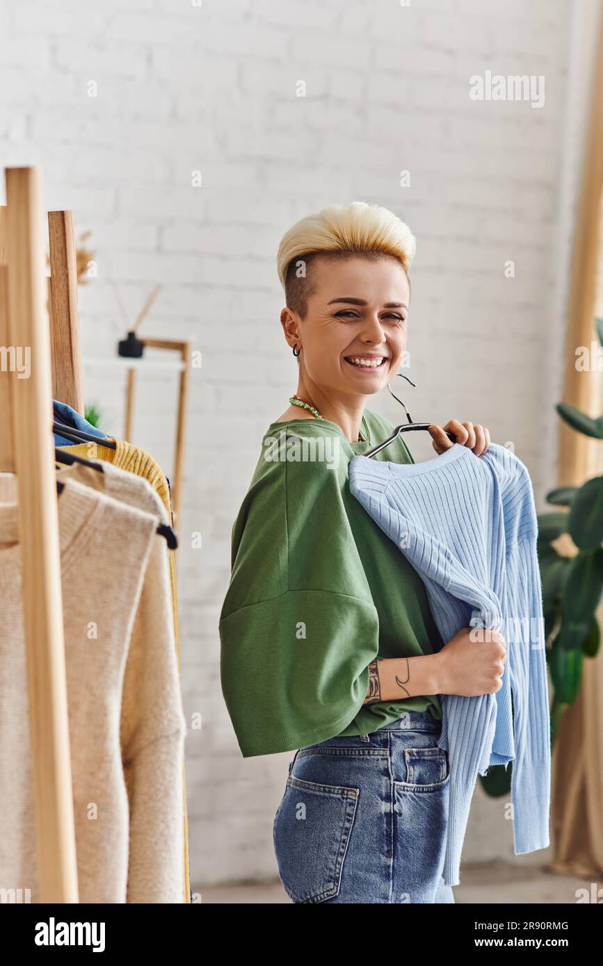 Aufgeregte tätowierte Frau mit trendigen Haaren, die eine blaue Strickjacke hält und in der Nähe eines Regals vor der Kamera lächelt, mit Second-Hand-Ladengegenständen auf Kleiderbügeln zu Hause, Sustain Stockfoto