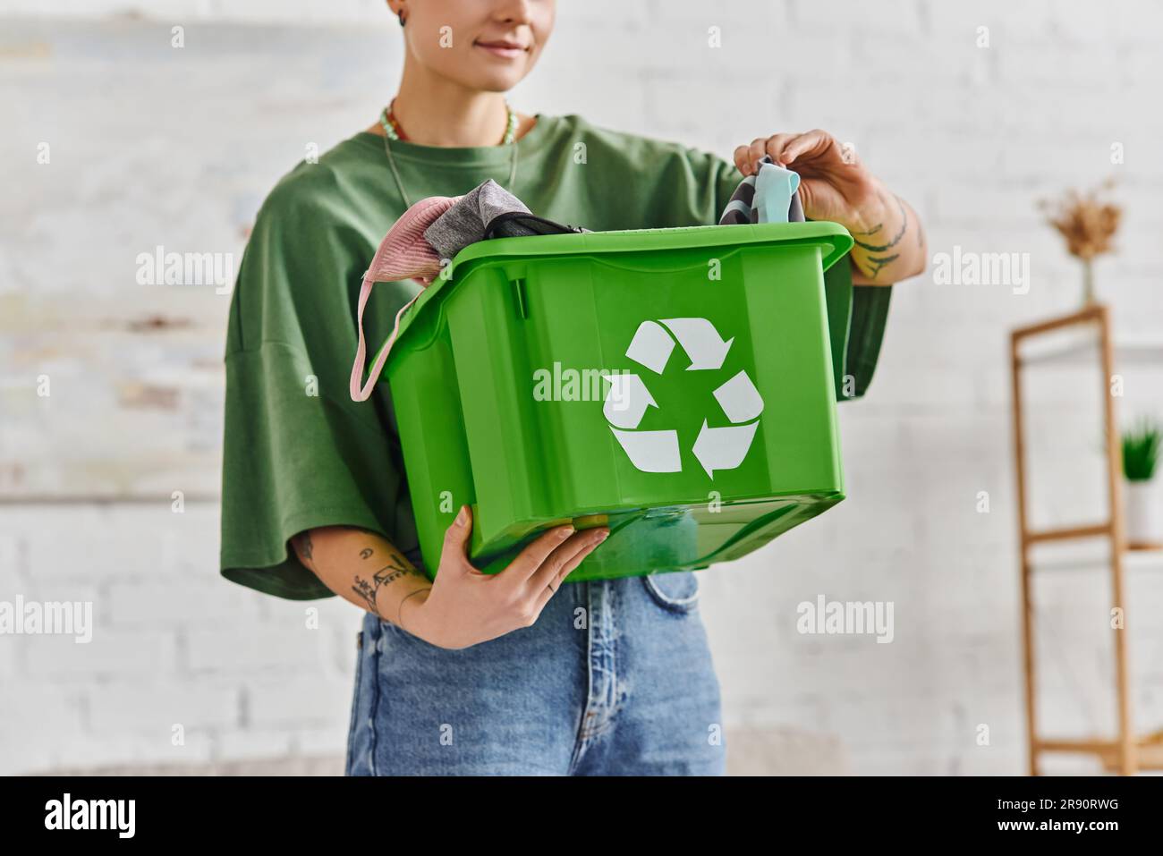 Teilansicht einer tätowierten Frau mit grüner Recyclingbox und Kleidung, während sie im Wohnzimmer steht, ethischer Konsum, nachhaltiges Wohnen und Umwelt Stockfoto