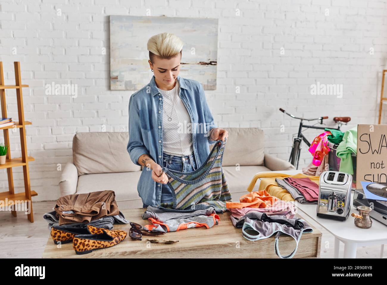 Junge tätowierte Frau, die ein gestricktes Tanktop neben Kleidungsstücken, einem elektrischen Toaster und einer Zauberei hält, während sie Liebesstücke für den Austausch zubereitete, sustai Stockfoto