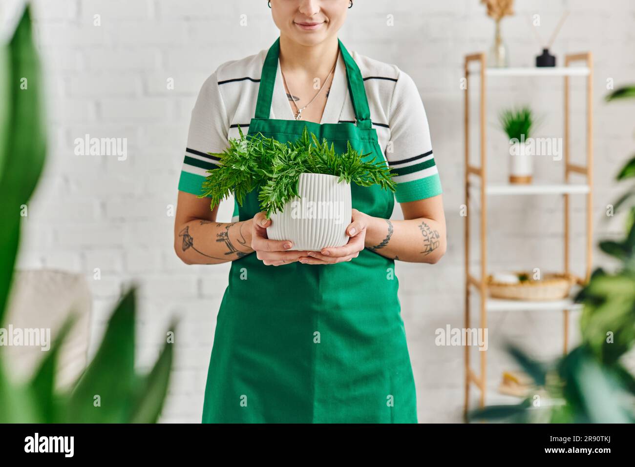 Nachhaltiges Zuhause, Innengärten, Teilansicht einer jungen tätowierten Frau in einer Schürze, die einen Blumentopf mit grüner Pflanze hält und in einer modernen Wohnung lächelt, Stockfoto
