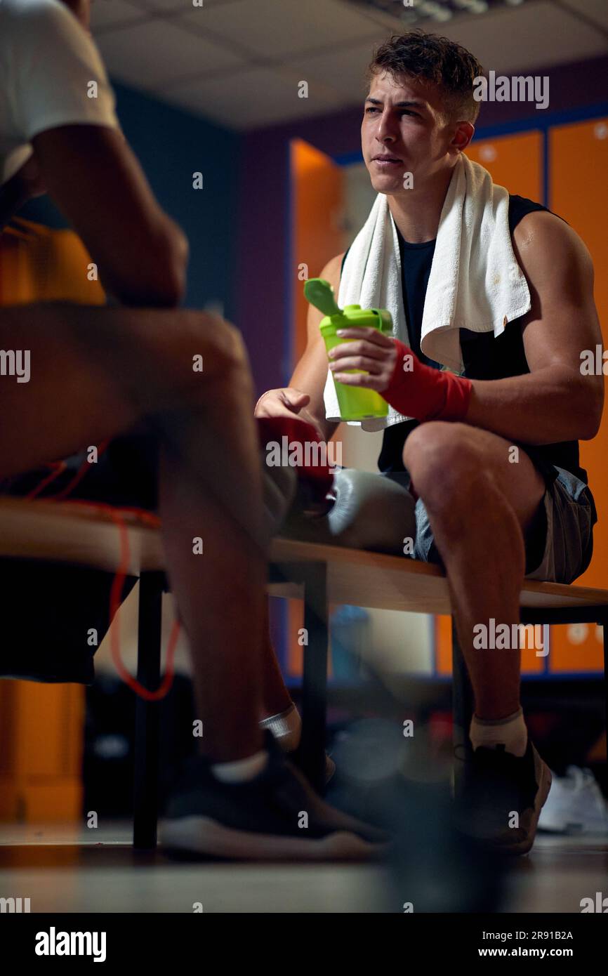 Junger muskulöser Boxspieler, der in der Garderobe sitzt, eine Wasserflasche hält, seinem Trainer zugewandt und spricht. Sport, Kampfsport, Lifestyle-Konzept. Stockfoto