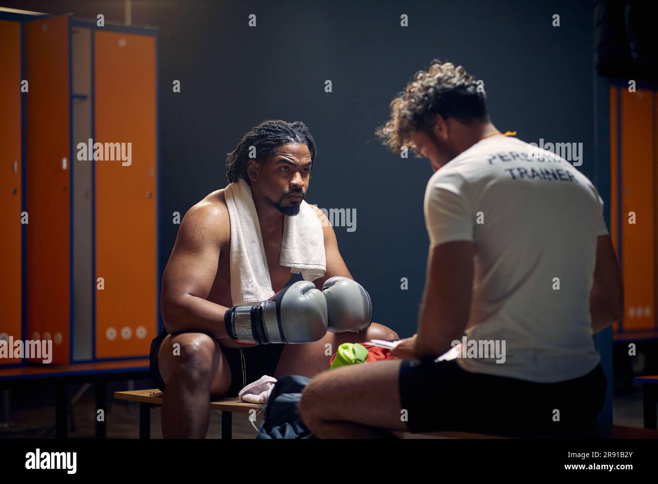 Breite Horizontalaufnahme eines starken, muskulösen Boxspielers nach dem Training in Schweiß, der mit seinem Trainer auf der Bank sitzt und spricht. Stockfoto