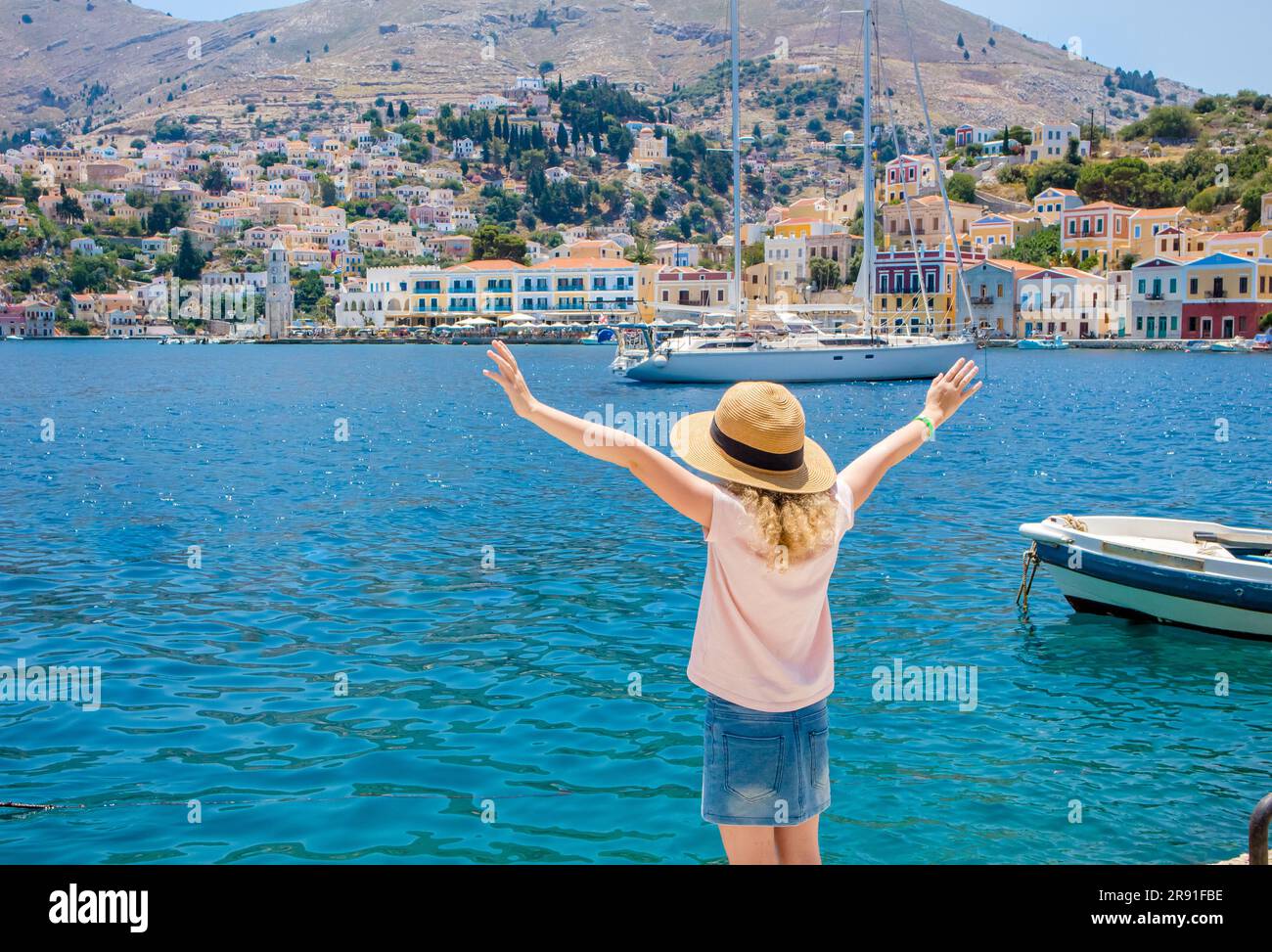 Süßes blondes Mädchen im Urlaub in Griechenland. Symi auch bekannt als Syme oder Simi, griechische Insel im Hintergrund. Symi Hauptstadt mit bunten Gebäuden. Stockfoto