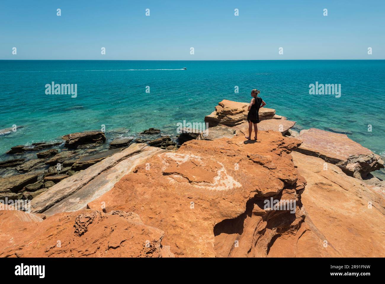 Eine Frau steht am Rand der Felsen am Gantheaume Point, während ein kleines Boot in Broome West Australia vorbeifährt Stockfoto