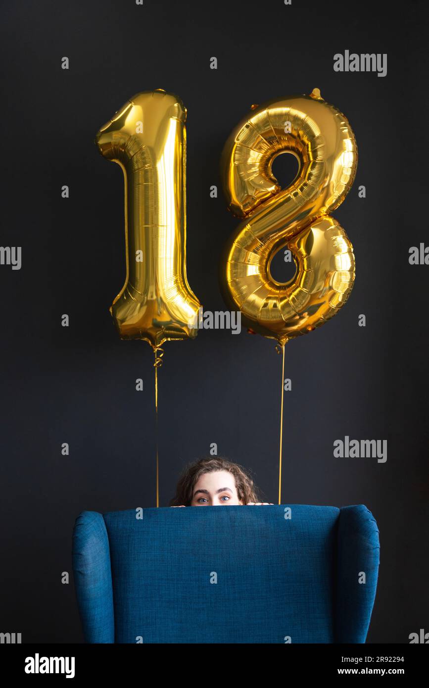 Eine Frau versteckt sich hinter einem Stuhl mit Ballon Nummer 18 auf schwarzem Hintergrund Stockfoto