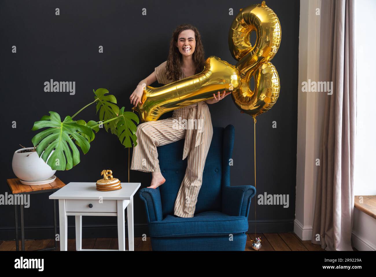 Verspielte junge Frau, die zu Hause auf einem Stuhl mit Heliumballon steht Stockfoto