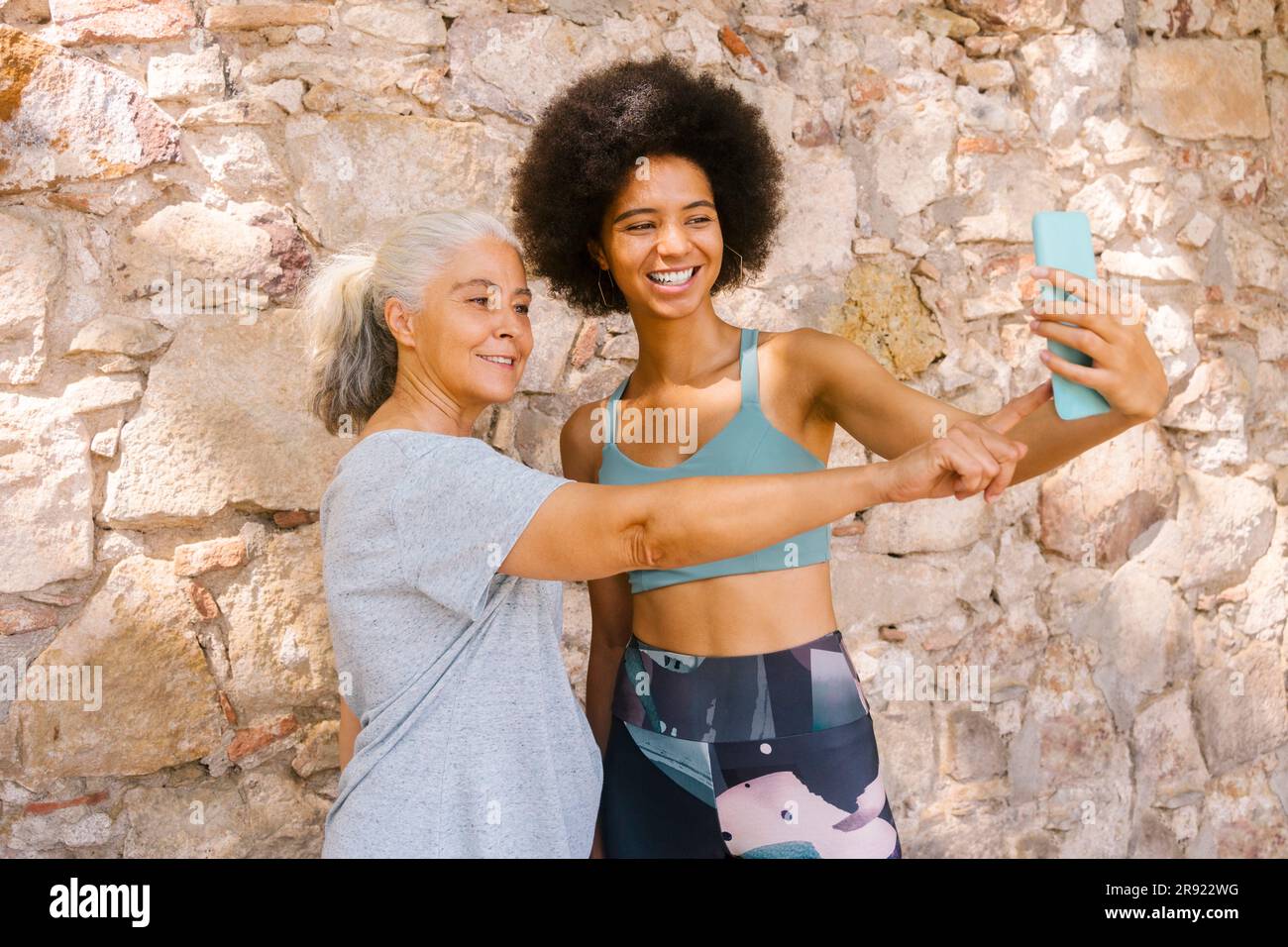 Lächelnde Frau, die Selfie mit Mutter vor einer Steinmauer macht Stockfoto