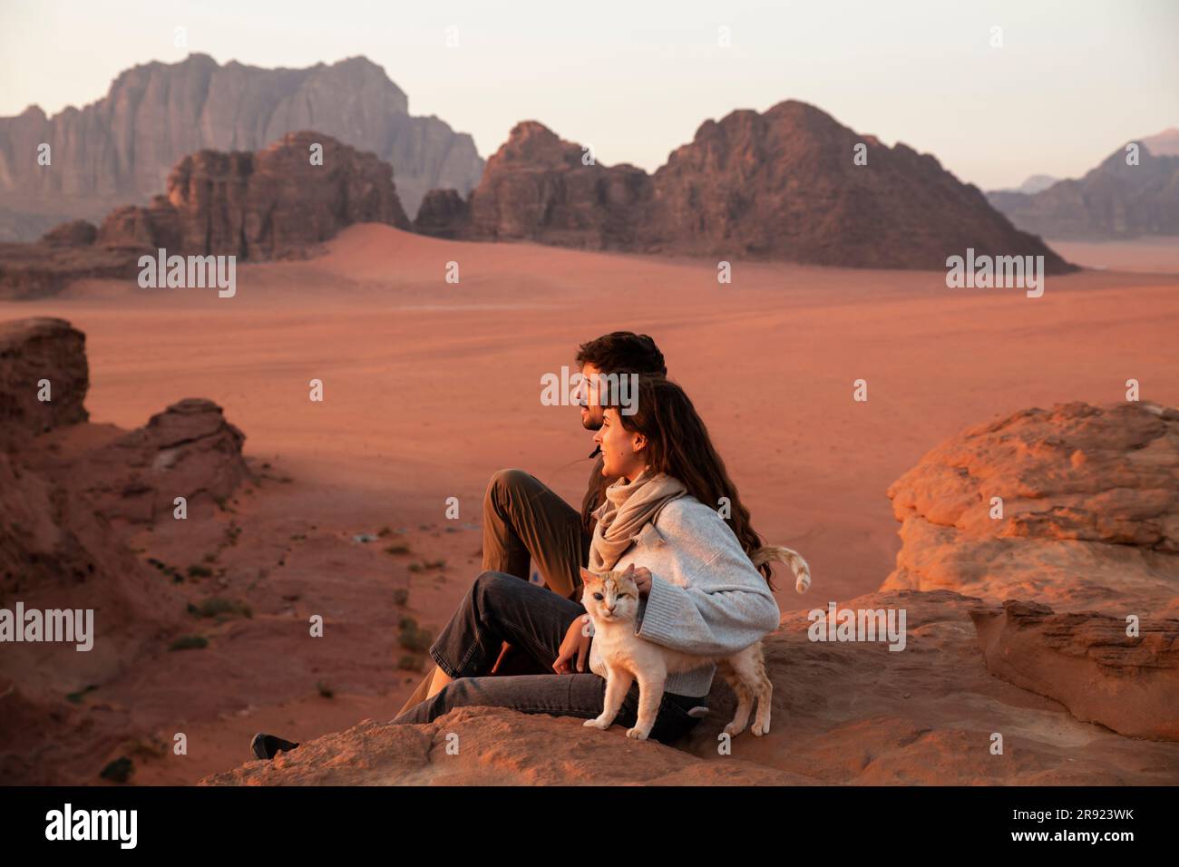 Ein junges Paar, das bei Sonnenuntergang mit der Katze auf Wüstenfelsen sitzt Stockfoto