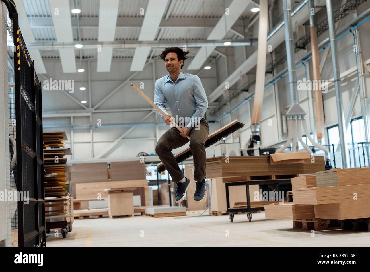 Glücklicher junger Geschäftsmann, der in der Industrie mit dem Besen springt Stockfoto