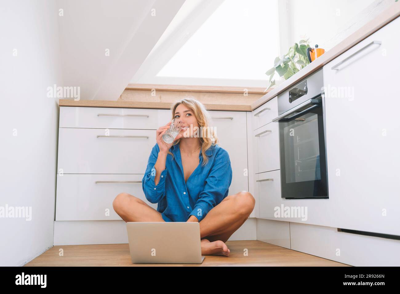 Lächelnde Frau, die Wasser trinkt und mit einem Laptop in der Küche sitzt Stockfoto