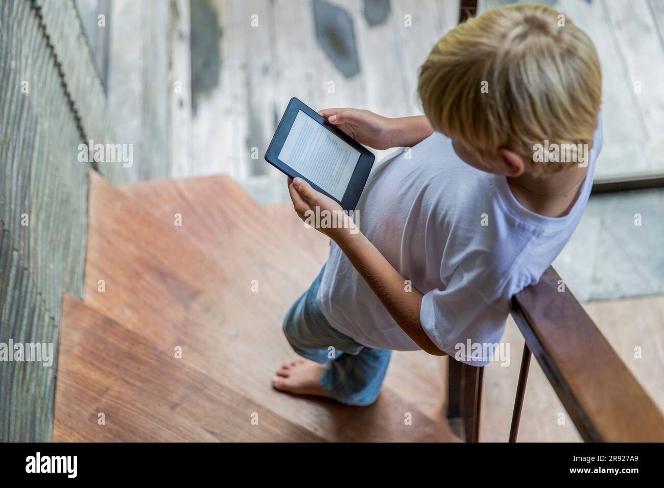 Ein blonder Junge, der E-Reader benutzt und zu Hause auf der Treppe steht Stockfoto