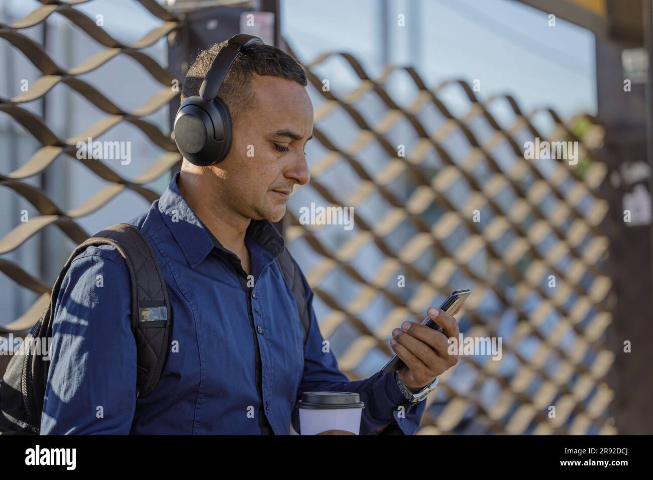 Mittellange Aufnahme eines jungen Latinos mit Kopfhörern, der an der Bushaltestelle von hinten auf sein Handy schaut. Stockfoto