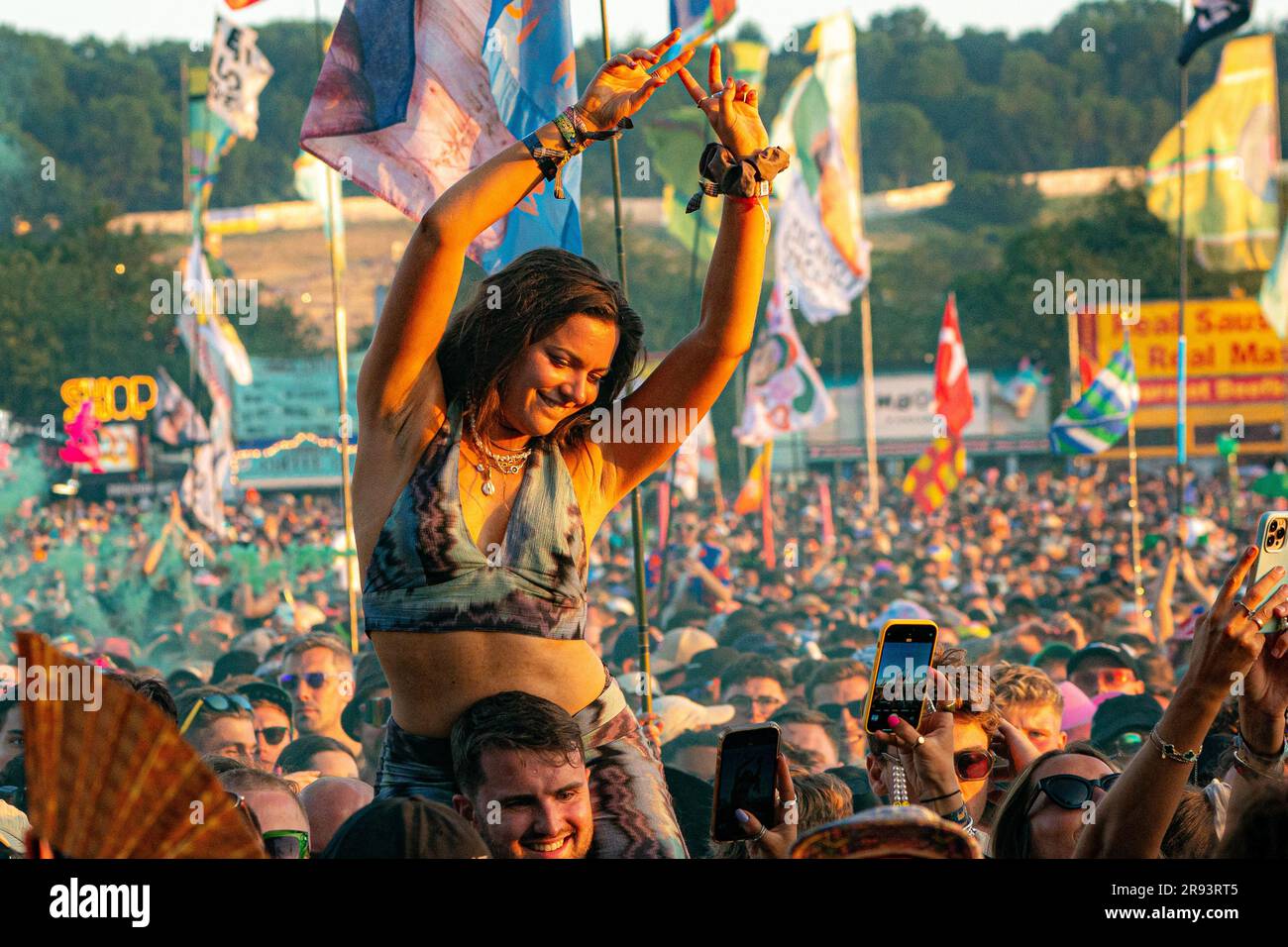Eine Frau lässt sich von einer Freundin fotografieren, während sie auf den Schultern tanzt und Fred auf der anderen Bühne beim Sonnenuntergang über dem Glastonbury Festival in Worthy Farm in Somerset zusieht. Foto: Freitag, 23. Juni 2023. Stockfoto