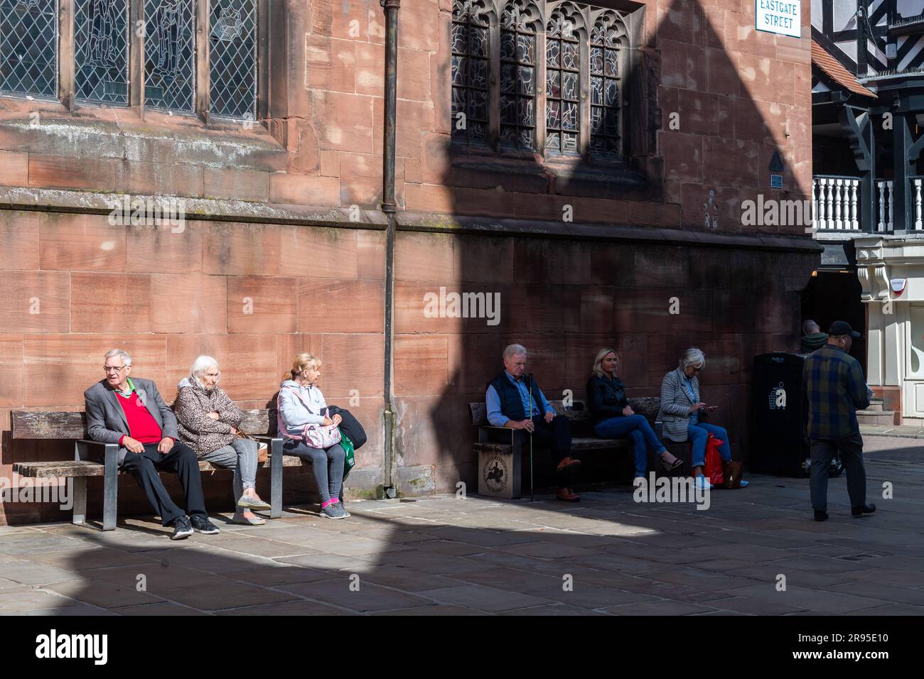 Leute, die auf Bänken im Sonnenlicht und im Schatten sitzen, Chester, Cheshire, Großbritannien. Stockfoto