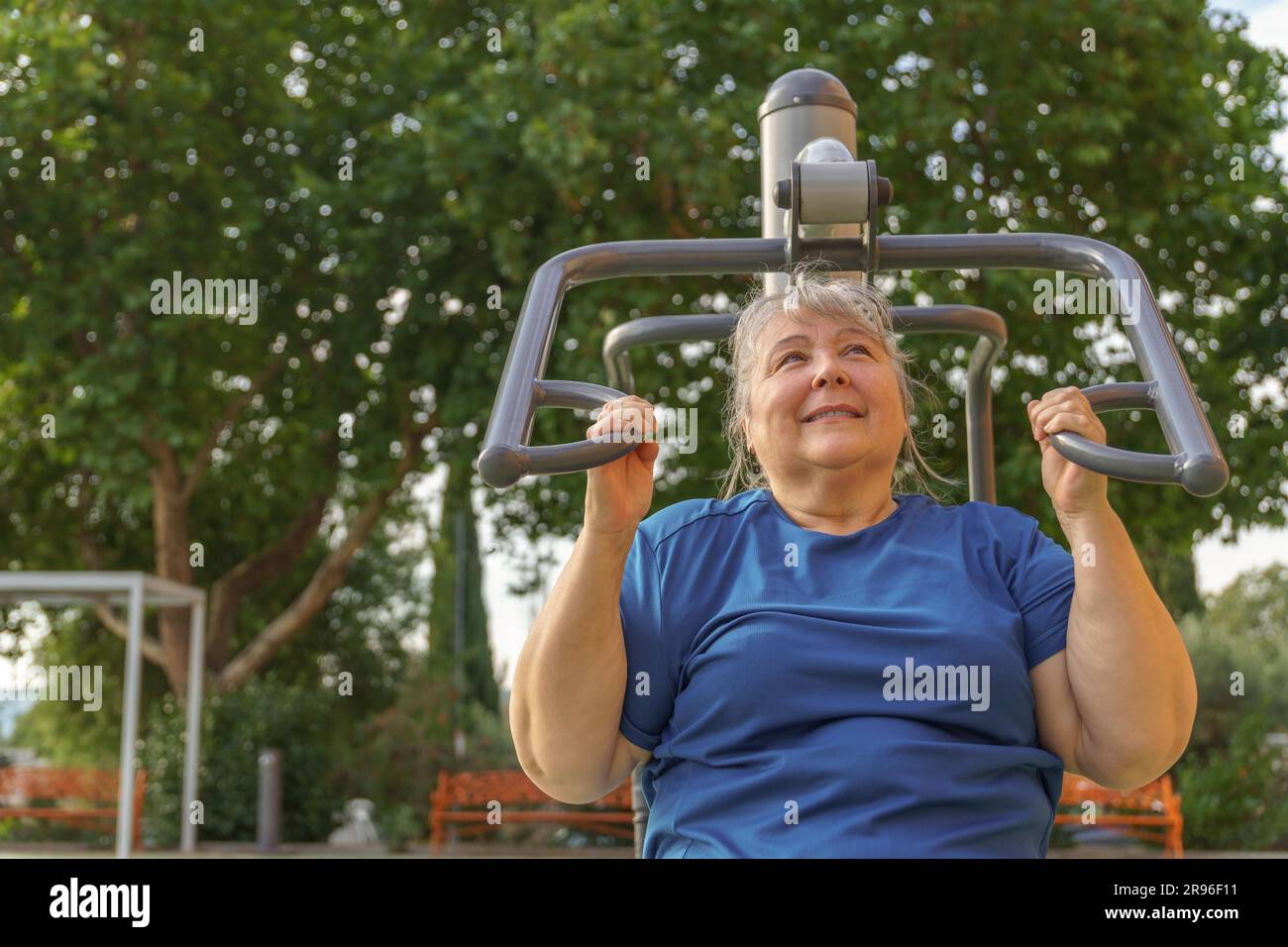 Ältere fettleibige Frau in Sportbekleidung, die in einem Outdoor-Fitnessstudio in einem Park, umgeben von Bäumen, Trainingsgerät, gesundem Lebensstil und schlankem Design trainiert Stockfoto
