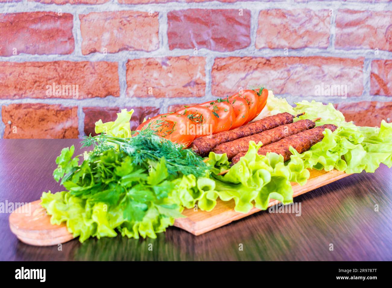 Lulya-Kebab. Shish Kebab auf einem Stock, Füllung. Traditionelles kaukasisches Gericht. Auf einem Schneidebrett mit grünem Salat, Ketchup, Gewürzen, Tomaten auf einem Ziegelstein Stockfoto