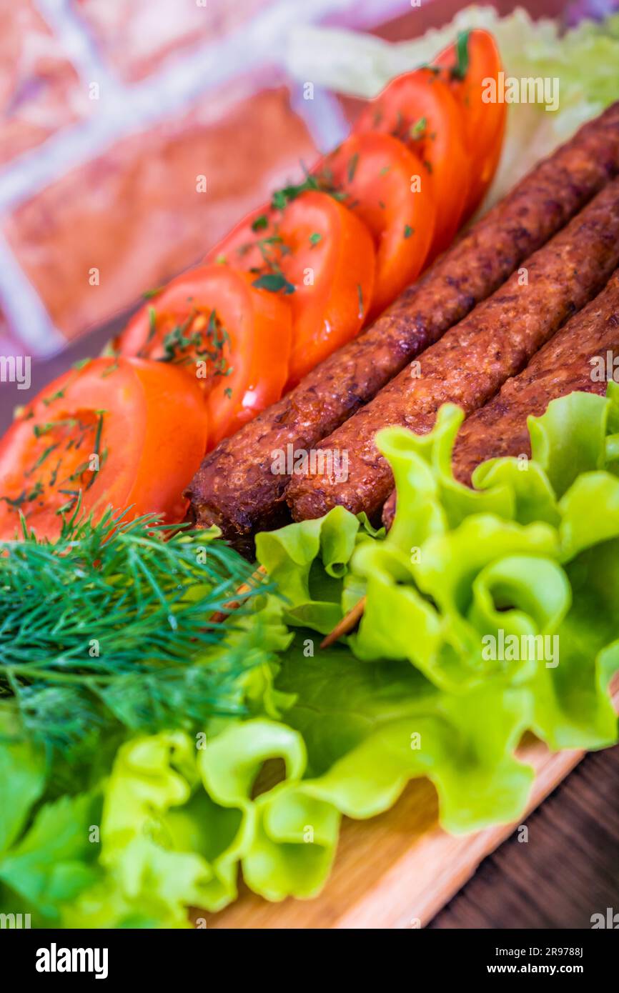 Lulya-Kebab. Shish Kebab auf einem Stock, Füllung. Traditionelles kaukasisches Gericht. Auf einem Schneidebrett mit grünem Salat, Ketchup, Gewürzen, Tomaten auf einem Ziegelstein Stockfoto