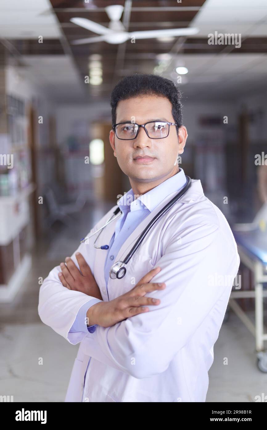 Porträt eines jungen Arztes im weißen Labormantel mit überkreuzten Armen. Glücklicher Seniorarzt im Mantel lächelnd auf Krankenhauskorridorhintergrund. Stockfoto