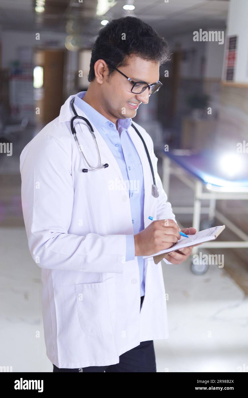 Porträt eines männlichen Oberarztes, der Notizen in Not Pad schreibt. Glücklicher indischer Arzt lächelnd, mit weißem Mantel im Krankenhausflur stehend. Stockfoto