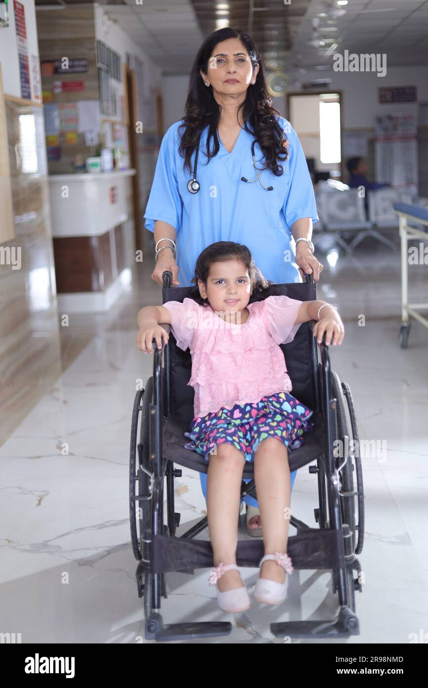 Kleines Mädchen Kind, das auf einem Rollstuhl sitzt und eine Krankenschwester sich um sie kümmert, beide stehen im Krankenhausflur, Verletzung, körperliche Behinderung. Stockfoto