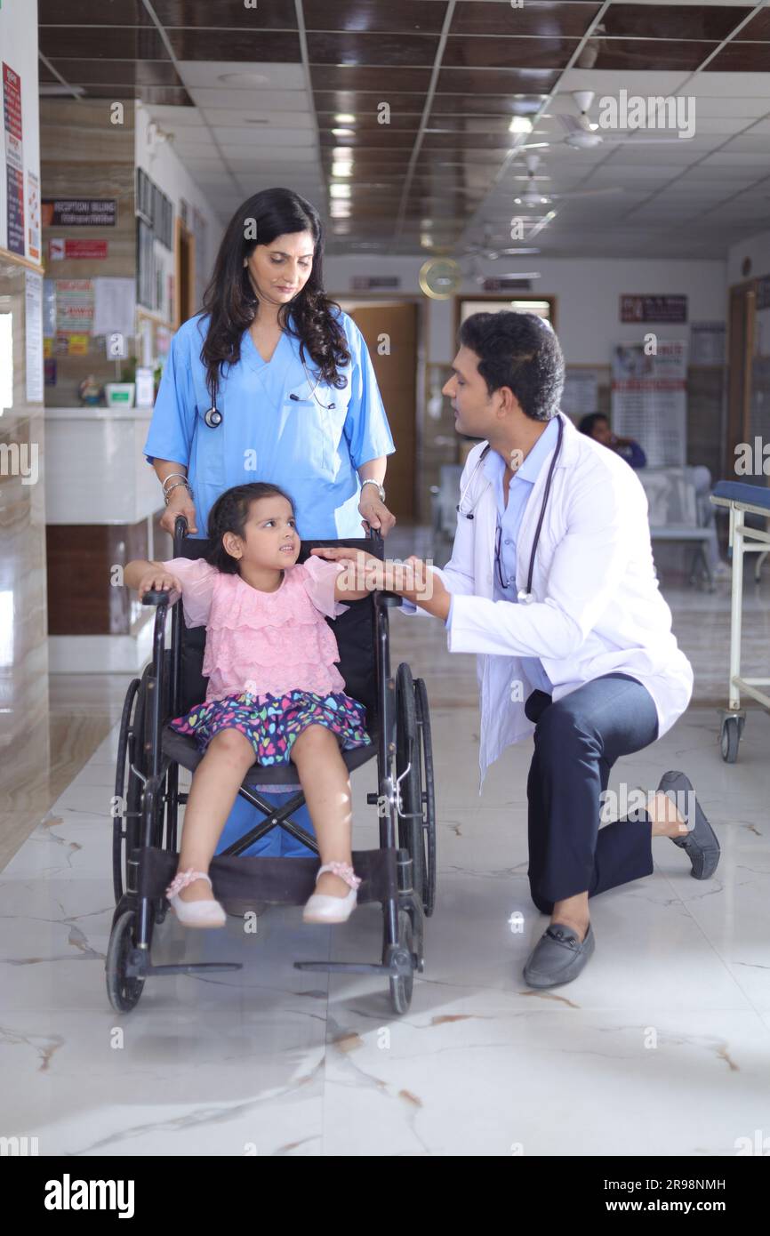 Mädchen Kind, das auf einem Rollstuhl sitzt, mit einer Krankenschwester und einem Arzt, die sich um sie kümmern, beide stehen im Krankenhausflur, Verletzung, körperliche Behinderung. Stockfoto