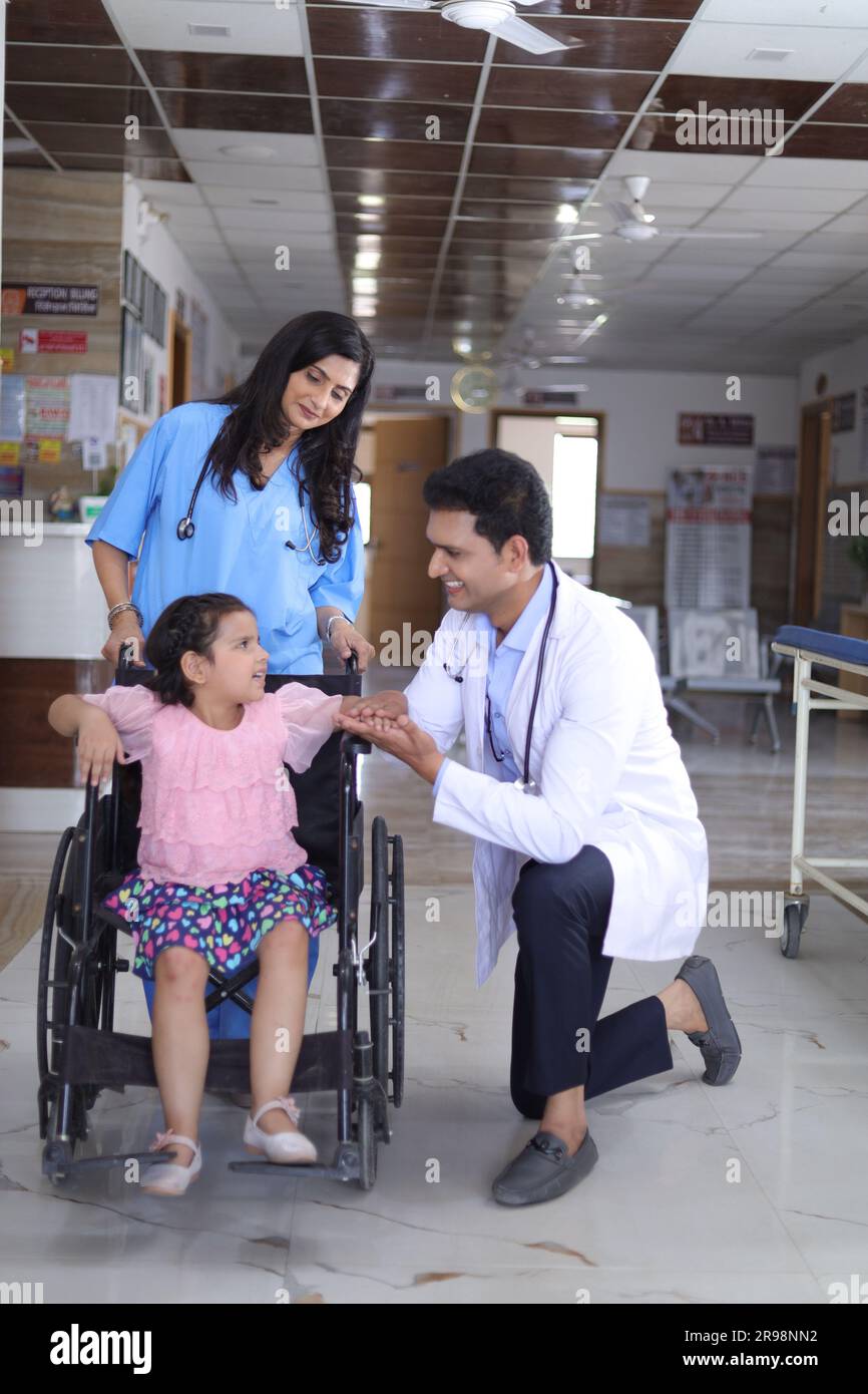 Mädchen Kind, das auf einem Rollstuhl sitzt, mit einer Krankenschwester und einem Arzt, die sich um sie kümmern, beide stehen im Krankenhausflur, Verletzung, körperliche Behinderung. Stockfoto