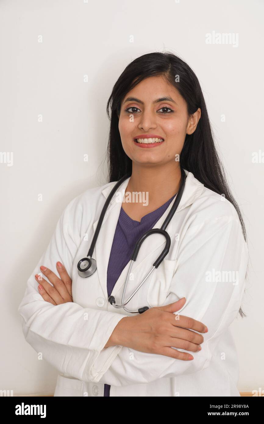 Porträt einer indischen lächelnden Ärztin im Laborkittel mit gekreuzten Armen vor blauem Hintergrund. Glücklicher indischer Arzt im weißen Mantel lächelnd auf Ho Stockfoto