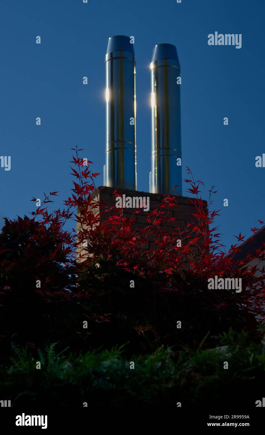 Rote Ahornblätter vor zwei modernen Stahlschornsteinen, die in hellem Sonnenlicht leuchten Stockfoto