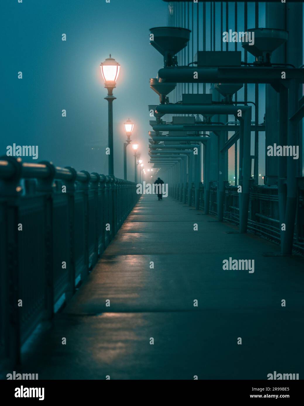 Ein Mann läuft über eine Brücke in einer nebligen Nacht, mit einem Lampenpfosten im Vordergrund Stockfoto