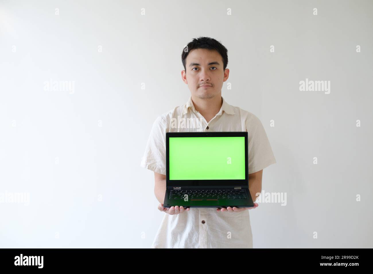 Ein asiatischer Mann mit beigefarbenem Hemd hält einen Laptop mit grünem Bildschirm mit beiden Händen und zeigt ihn der Kamera Stockfoto