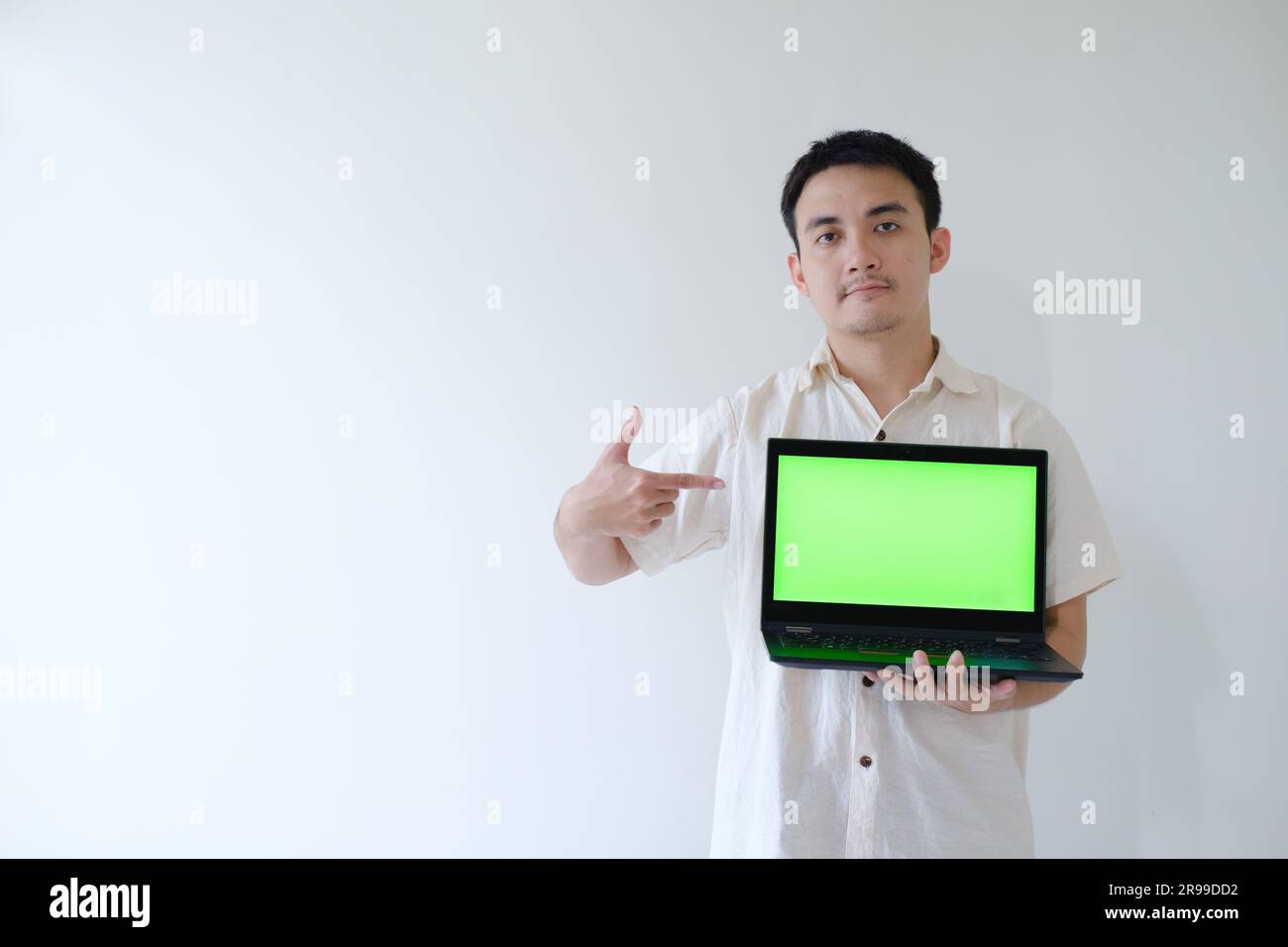 Ein asiatischer Mann, der ein graues Hemd trägt, zeigt mit seiner rechten Hand auf einen Laptop mit einem grünen Bildschirm in der linken Hand. Ich zeige es der Kamera. Stockfoto