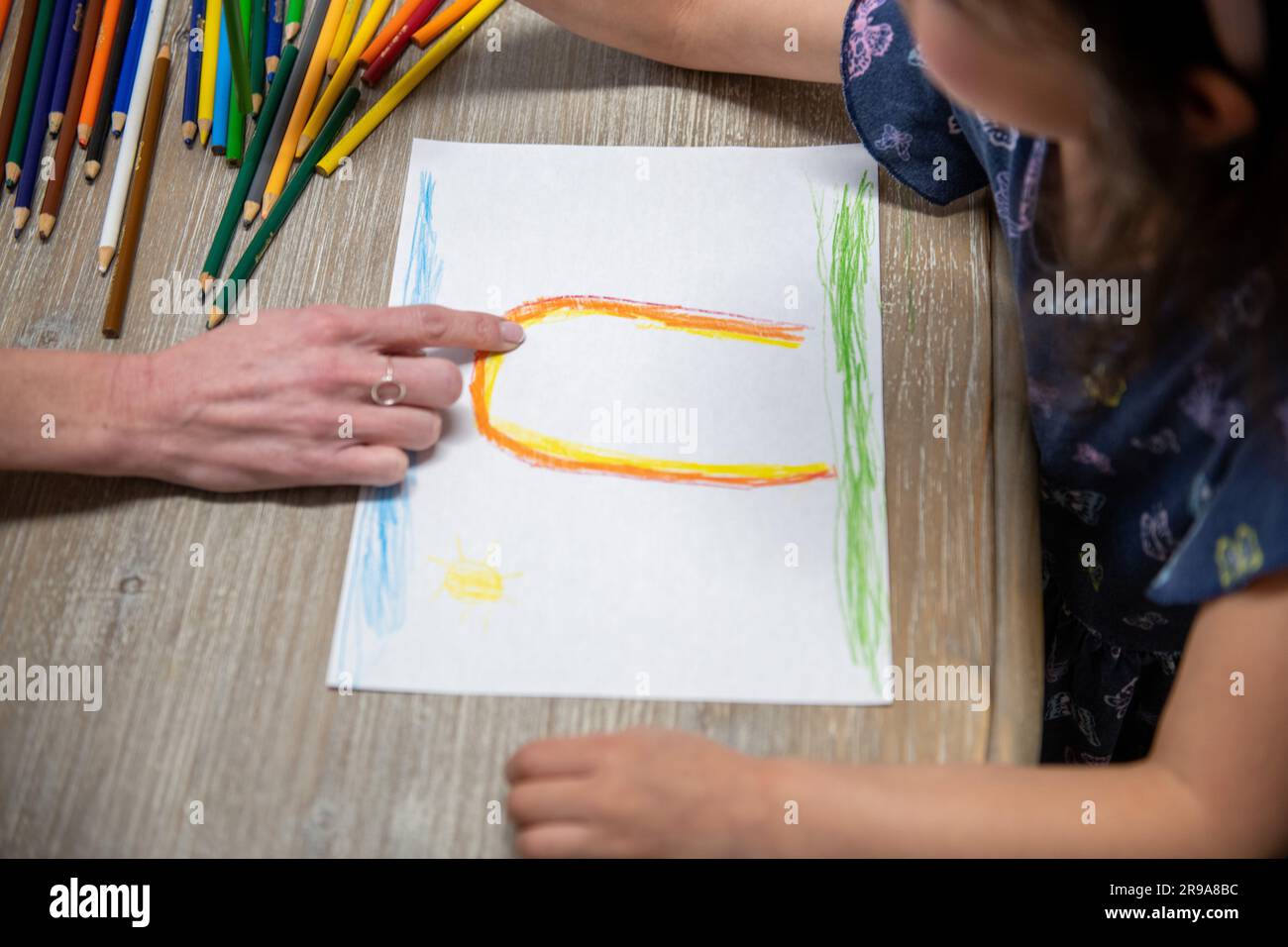 Ein kleines Kind zeichnet auf einem Stück Papier, während eine Erwachsene Hand auf das Kunstwerk zeigt Stockfoto