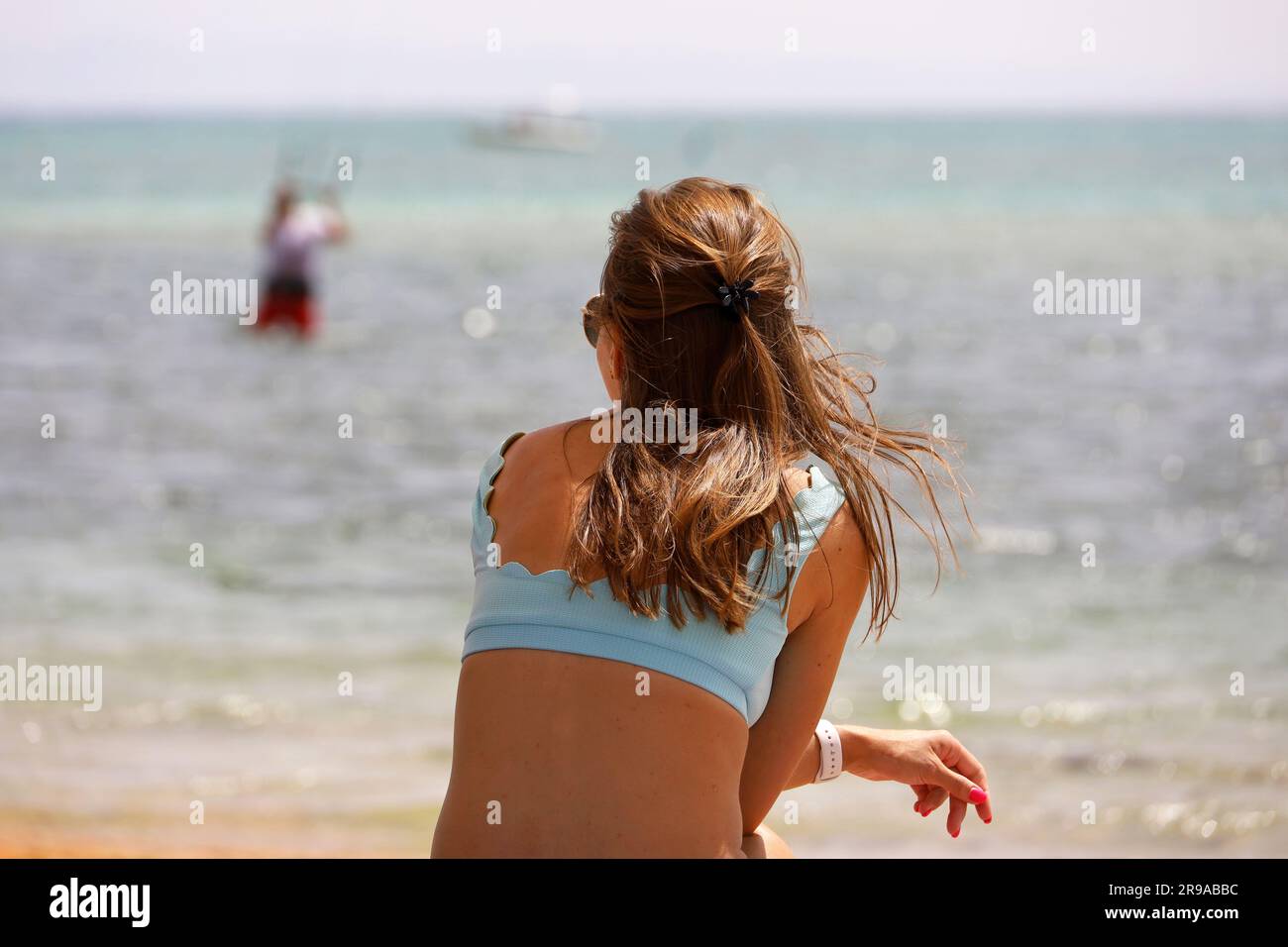 Ein Mädchen im Badeanzug sitzt am Strand und sieht einen Mann an, der Parasailing betreibt. Entspannung im Sommerresort Stockfoto
