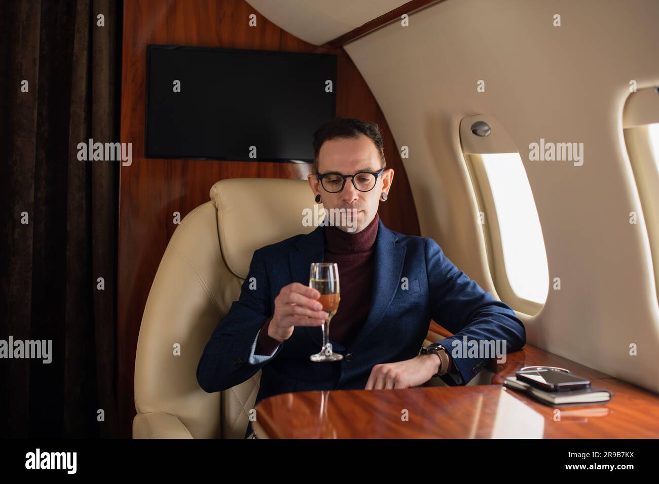 Eleganter junger Geschäftsführer mit Brille in Jacke sitzt in einem privaten Flugzeug Jet und trinkt Champagner während des Fluges Stockfoto