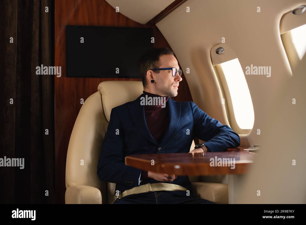 Junger, eleganter, selbstbewusster männlicher Geschäftsmann mit Brille in blauer Jacke, der durch ein Firmenflugzeugfenster blickt, Geschäftskonzept für Reisen und Fluggesellschaften Stockfoto